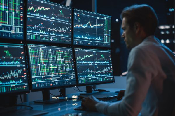 Financial Analyst Working on a Computer with Multi-Monitor Workstation with Real-Time Stocks, Commodities and Exchange Market Charts. Businessman Works in Investment Bank Downtown Office at Night.